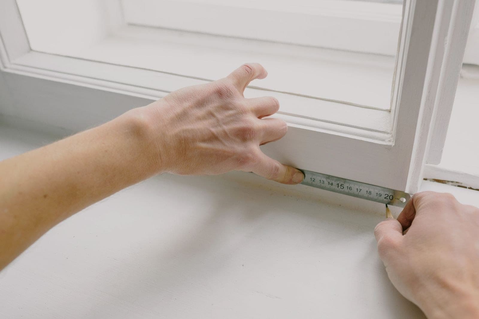 Hands measuring a white window frame with a steel ruler and pencil