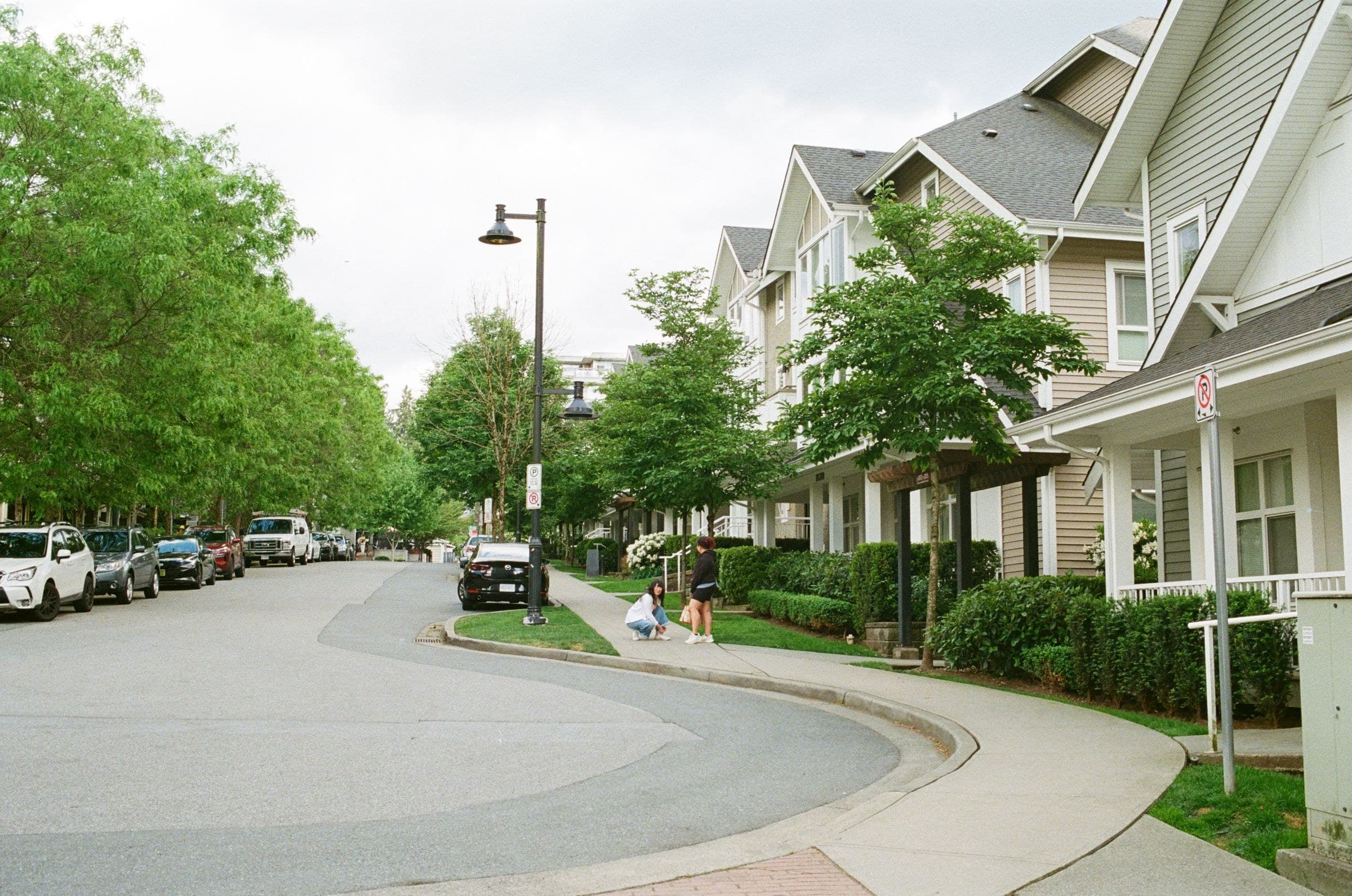 Curving residential street of two-storey houses with siding, street trees and parked cars